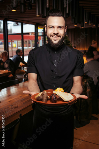 Cheerful bearded waiter with kebab and side dish in a georgian restaurant. A discerning waiter in black clothes, with a beard and a portion of kebabs