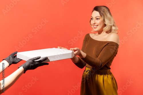 Woman taking pizza from courier hands in protective gloves, safe food delivery on quarantine, coronavirus 2019-ncov prevention. Stay home, order online. indoor studio shot isolated on red background