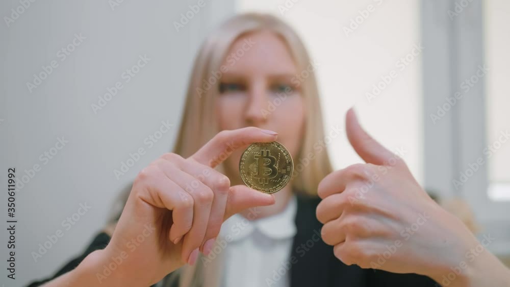 Smiling cheerful blond woman in office suit sitting at workplace with computer and showing bitcoin in hand doing thumbs up gesture and looking at camera Smiling cheerful blond woman in office suit sitting at workplace with computer and showing bitcoin in hand doing thumbs up gesture and looking at camera