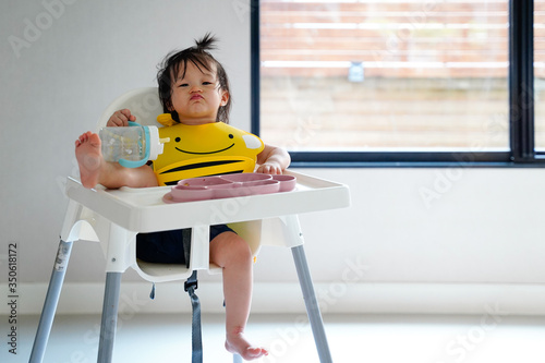 Asian naughty baby girl 1 year old eating on the high chair and holding water bottle by her hand with wearing yellow apron, a foot on a table with cute posture at home next window.