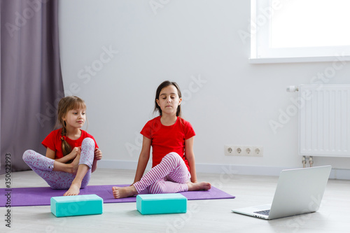 two little girls practicing yoga, stretching, fitness by video on notebook. Distant online education training, aerobic at home. Healthy lifestyle, coronavirus, stay home. Kids sport home quarantine.