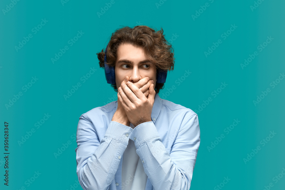 Blurt out. Portrait of a young beautiful man wearing white t-shirt and blue shirt in headphones closes his mouth with his hands because he blurted something out Blurt out. Portrait of a young beautiful man wearing white t-shirt and blue shirt in headphones closes his mouth with his hands because he blurted something out