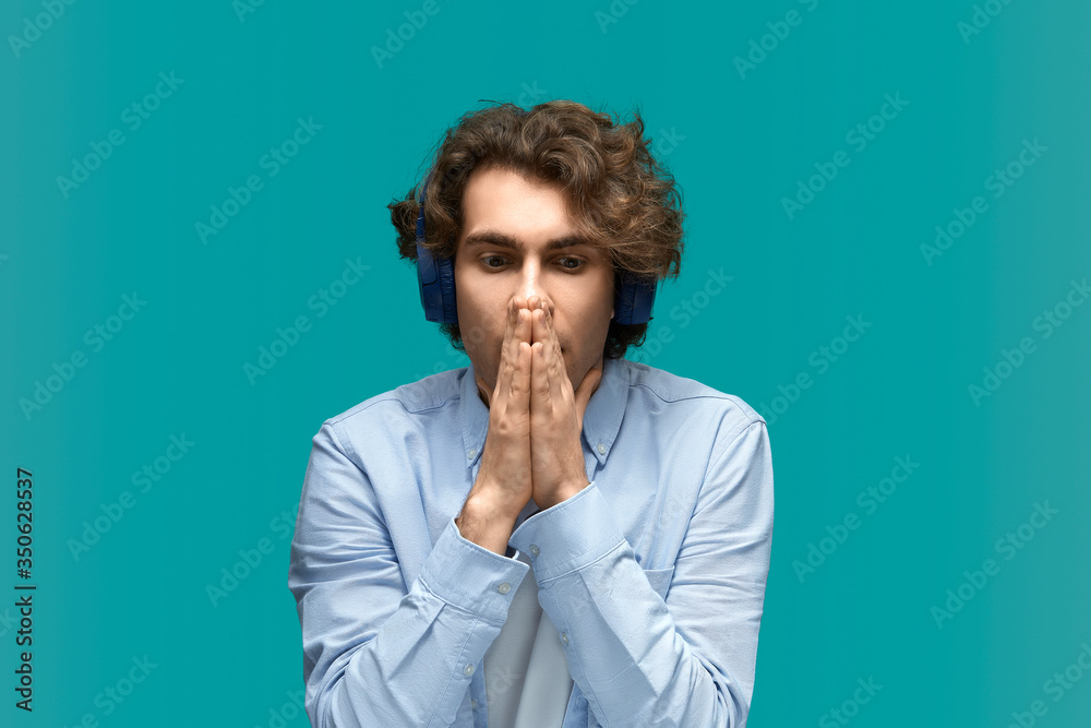 In trouble. Portrait of a young beautiful man wearing white t-shirt and blue shirt in headphones closes his mouth by boat hands