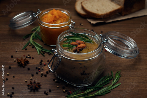 Glass jar with poultry pate, decorated with almonds and rosemary with orange jam on a wooden table