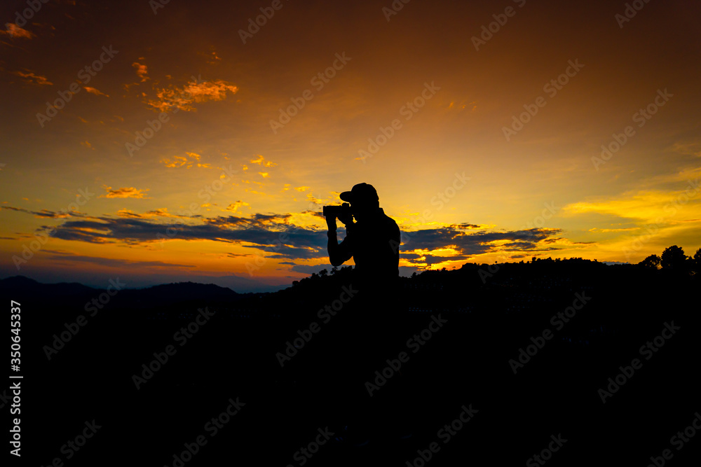 Silhouette Man Standing on Hill with  Camera at the Sunset on Mountain with Orange Sky. Enjoying Peaceful Moment Concept.