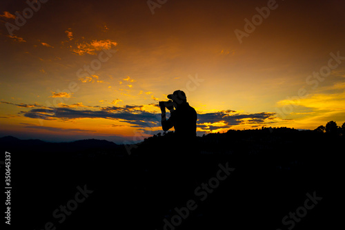 Silhouette Man Standing on Hill with Camera at the Sunset on Mountain with Orange Sky. Enjoying Peaceful Moment Concept.