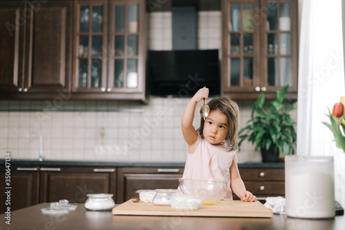 Cute little girl looks at the raised spoon she used to beat the eggs with for the dough at the table in kitchen with modern interior.
