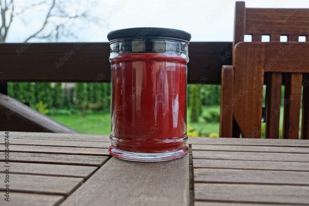 one red glass jar with a decorative candle stands on a brown board on the veranda against a background of green vegetation one red glass jar with a decorative candle stands on a brown board on the veranda against a background of green vegetation
