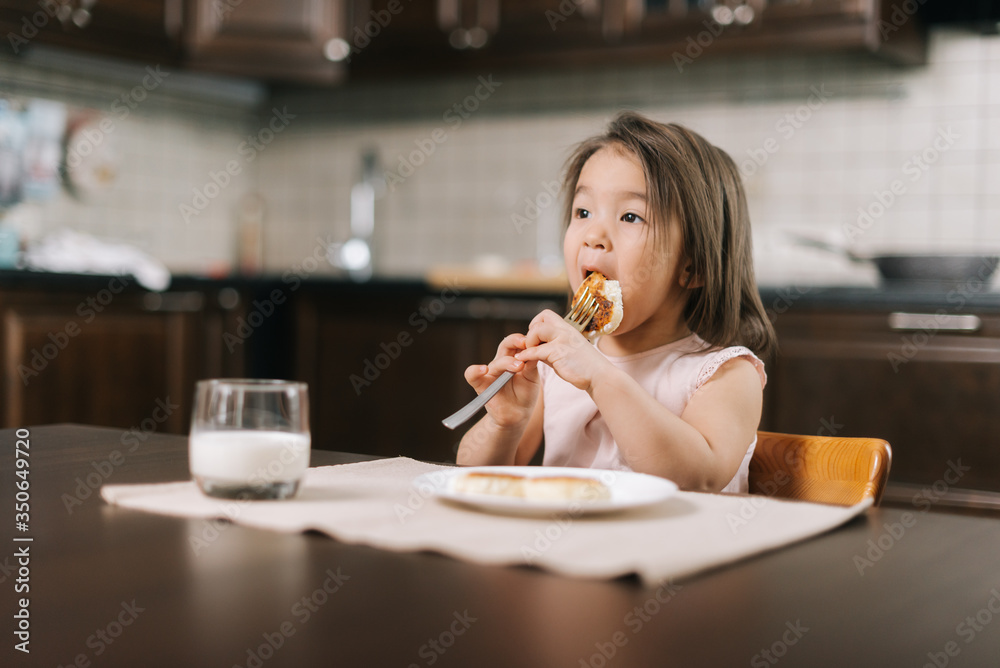 Hungry beautiful little two-year-old girl wearing white dress is eating tasty cheesecake with fork for lunch at the table in kitchen room. Glass of milk on the table. Hungry beautiful little two-year-old girl wearing white dress is eating tasty cheesecake with fork for lunch at the table in kitchen room. Glass of milk on the table.