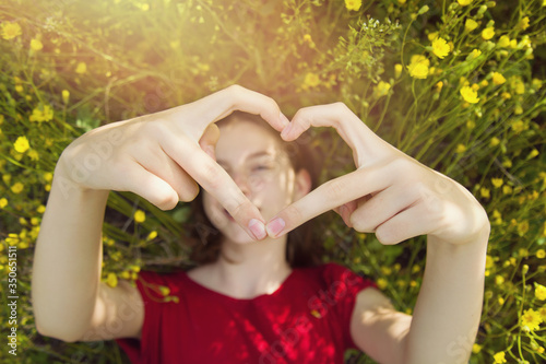 Happy young girl lies in the dense green grass in the park and shows hands a heart shape