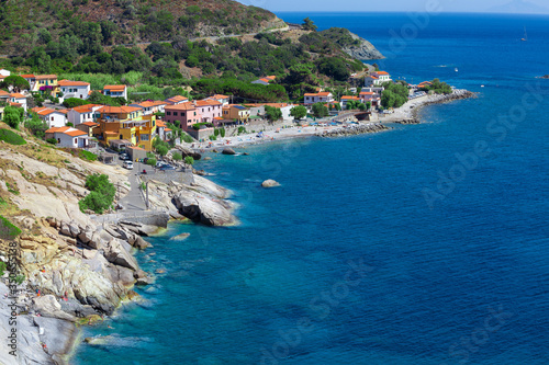 Sea shore with beach and rocks and rocky slope of the Island of Elba in Italy. Many people on the beach sunbathing. Blue sea with aerial view. Dwellings of a small village.