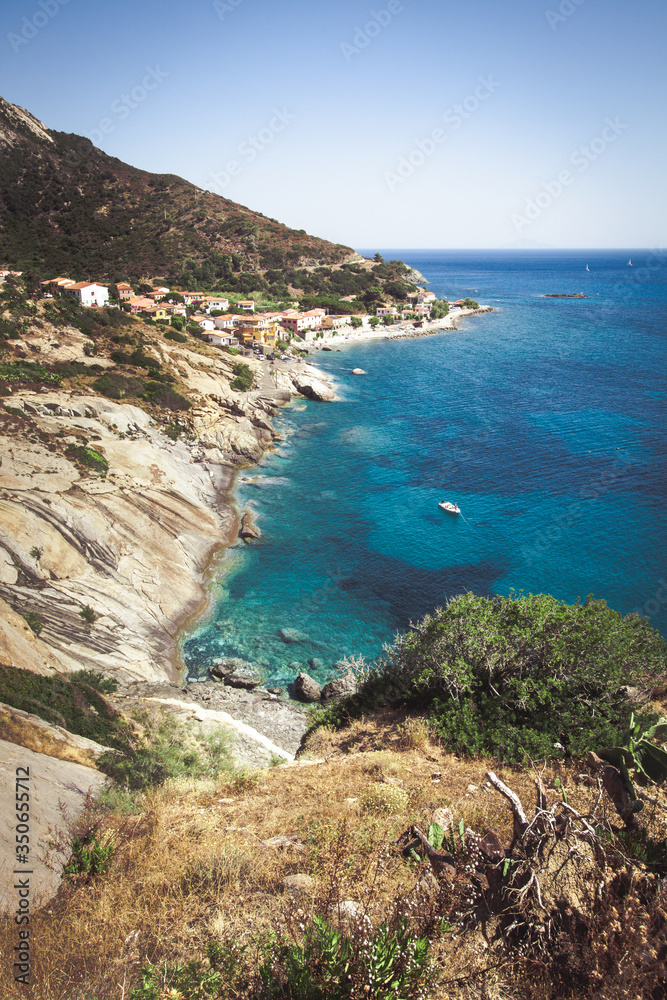 Seashore coastline with beach and rocks and rocky slope of the Island of Elba in Italy. Many people on the beach sunbathing. Blue sea with aerial view. Dwellings of a small village.