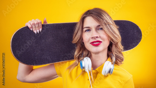 Happy lady in casual yellow polo with red lips and headphones holding skateboard on shoulder and looking at camera while standing on yellow background