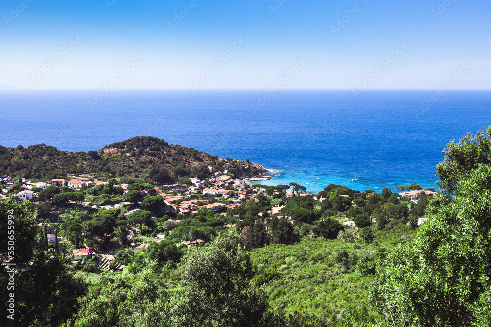 Seashore coastline with beach and rocks and rocky slope of the Island of Elba in Italy. Many people on the beach sunbathing. Blue sea with aerial view. Dwellings of a small village.