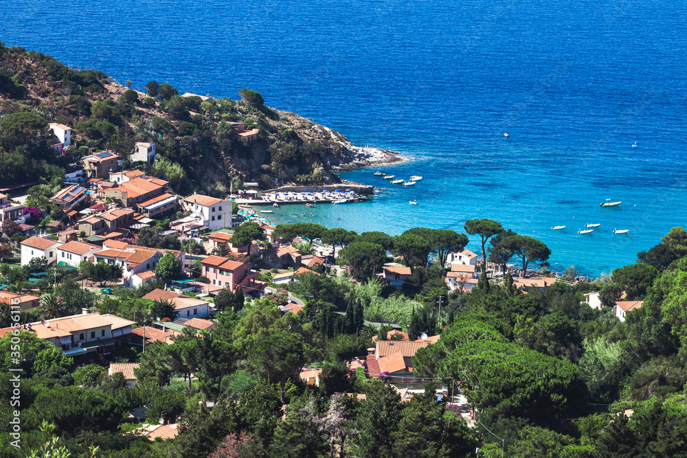 Fototapeta premium Seashore coastline with beach and little village in the Island of Elba in Italy. Many people on the beach sunbathing. Blue sea with aerial view. Houses in the middle of a natural landscape.