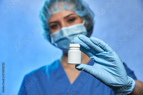 White bottle with pills on foreground in hand in glove of blurred doctor in medical mask and cap on background  Coronavirus COVID-19
