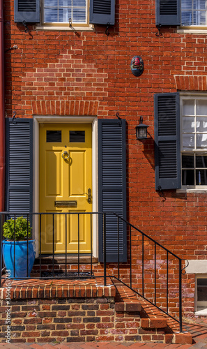 Old house in Annapolis Maryland with elegant yellow door with protective shutter, orange brick walls, steps leading to the door with black railings