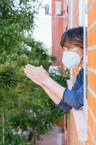 Woman clapping from her balcony to support those fighting the coronavirus. Masked woman clapping from her window. Applause from the window. Social distance. Older woman clapping. Coronavirus. © Tomaspf_