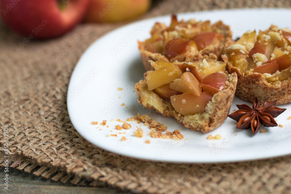 Crispy mini tartlets made from shortcrust pastry with apples, homemade sweets with anise and cinnamon. Serving on a white plate, on a wooden background. Natural eco sweets concept, warm cozy tea party