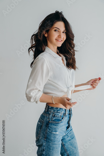 Smiling attractive young woman with dark hair in white shirt posing isolated on white wall background studio portrait. Life style concept.  Looks at the camera.