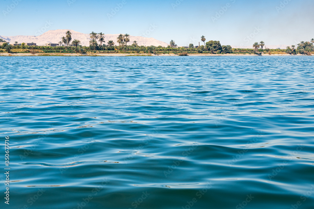 Fototapeta premium Surface of Nile river with palm trees on shore and chalk stone dunes at background