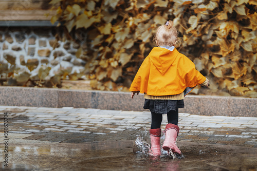 A joyful small girl in a yellow raincoat and pink rubber boots jumps on puddles with splashes and rejoices. Park, nature, outdoors. Autumn. Universal Children's Day. © Anna Shnaider
