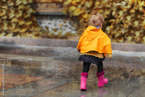 A happy little girl in a yellow raincoat and pink rubber boots walking in the rain on the park alone. Nature, outdoors. Childhood concept. Universal Children's Day.