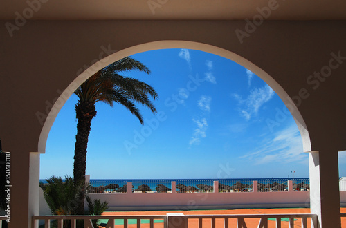 View of palm tree, deck chairs, beach and blue sky in clear weather from under the arch of the building