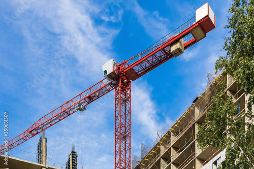 One high-rise crane against a house and sky during the construction phase. Industry concept for low-income young families. Mortgage, business, real estate loan. Summer. Green tree.