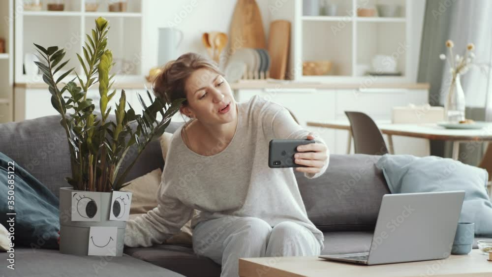 Young pretty woman sitting at table with tea mug, looking at camera, waving and telling about her houseplant in pot decorated with smiley face while chatting via video call at home under lockdown Young pretty woman sitting at table with tea mug, looking at camera, waving and telling about her houseplant in pot decorated with smiley face while chatting via video call at home under lockdown