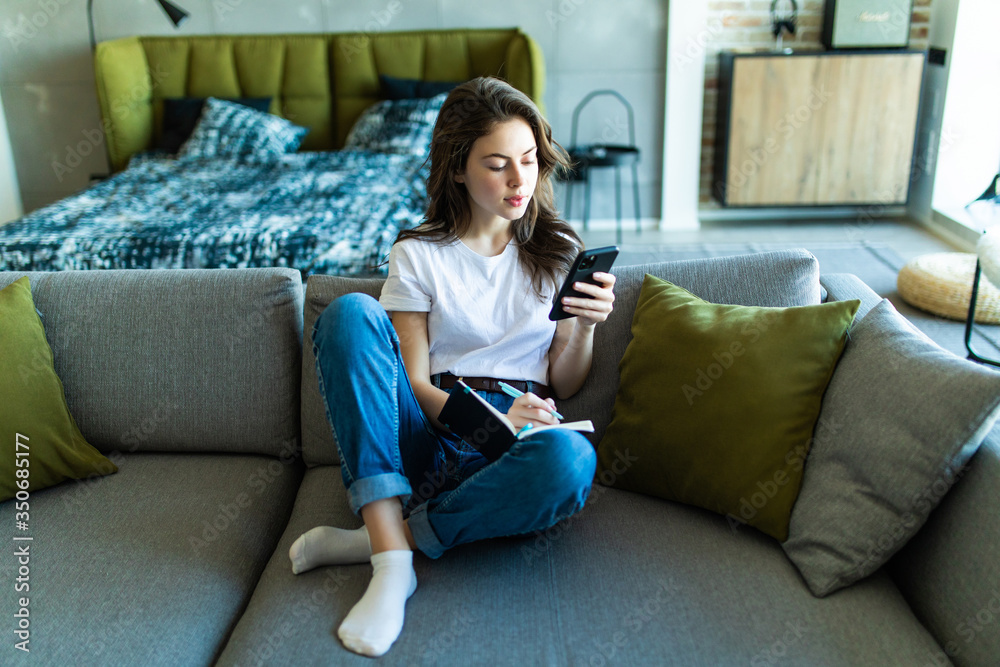 Smiling woman sitting near sofa, writing in weekly planner and surfing in phone. Working at home. Smiling woman sitting near sofa, writing in weekly planner and surfing in phone. Working at home.