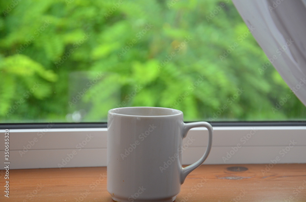 A white cup of tea or coffee stands on a wooden window sill against the background of a summer garden A white cup of tea or coffee stands on a wooden window sill against the background of a summer garden