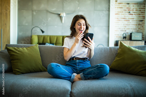 Portrait of a surprised woman holding a phone looking at you sitting on a sofa in the living room photo