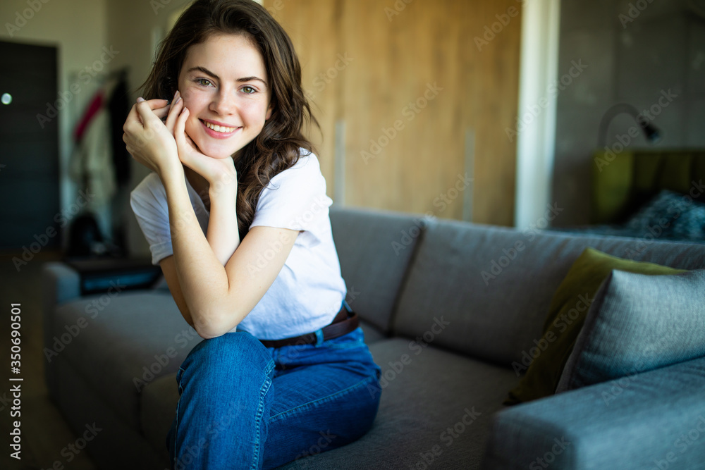Young dreaming woman sitting on sofa at home. Thoughtful young woman sitting in living room and looking up. Smiling woman relaxing on couch in a modern home.