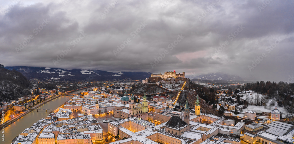 Panoramic aerial drone shot view of snowy Salzburg old town and Hohensalzb with city lights on during winter dusk hour
