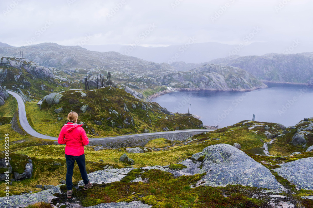 Tourist girl looks at the lake and the narrow road in the mountains at foggy cloudy weather in summer. On the way to Forsand. Norway.
