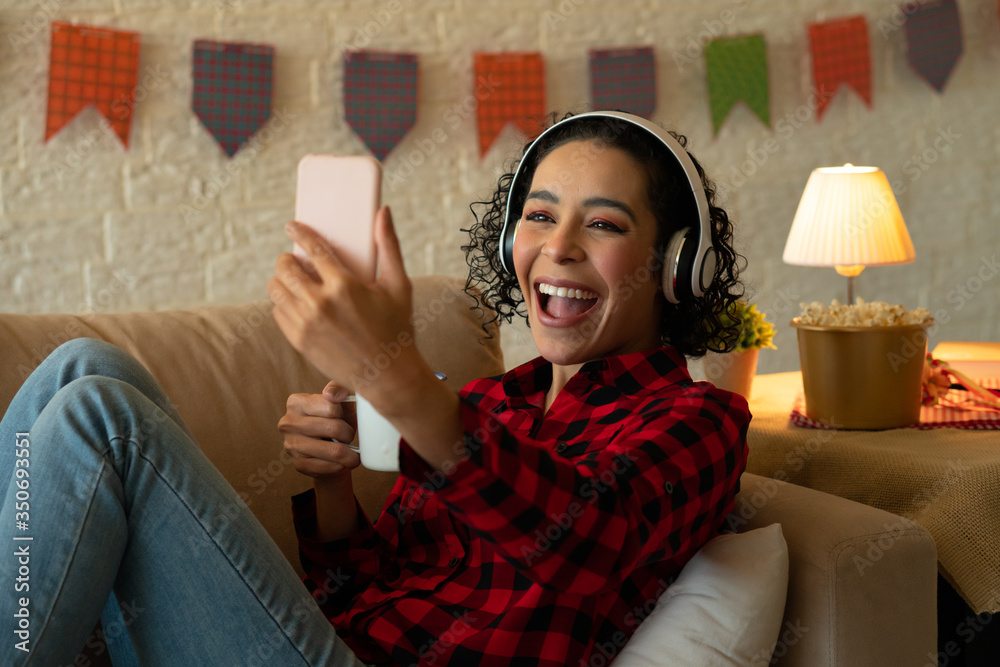 Cheerful african american woman with headphone smiling with broad smile and using mobile phone Indoors at home living room. Party, weekend, dance, quarantine, concert, music concept. - obrazy, fototapety, plakaty 