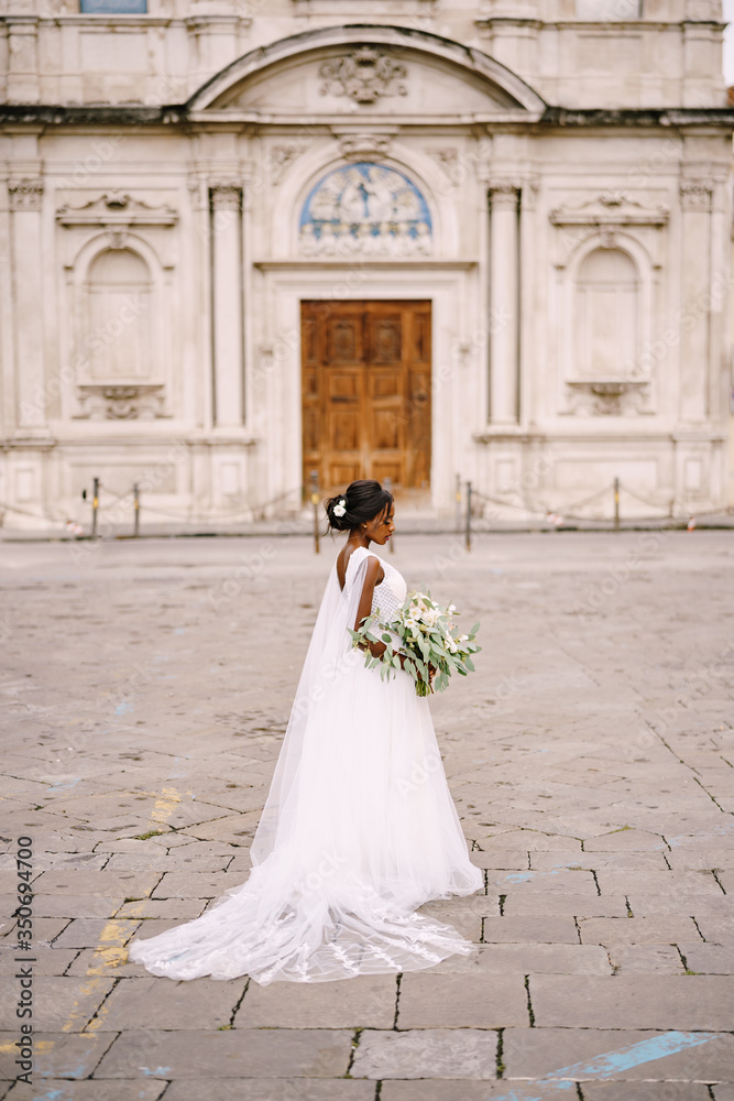 Interracial wedding couple. Wedding in Florence, Italy. African-American bride in a white dress with a bouquet in her hands, with a long veil stands on the street, against the background of the church