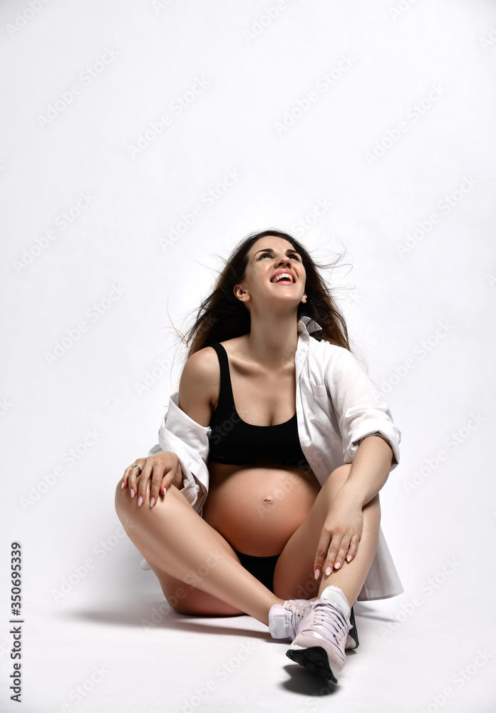 Obraz premium Pregnant lady in black sport shorts and top, shirt, pink sneakers. She is laughing, sitting on floor isolated on white. Close up