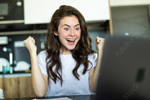 Free Download Excited young woman with win gesture with fist up sitting at table and looking at laptop - GFXLoad