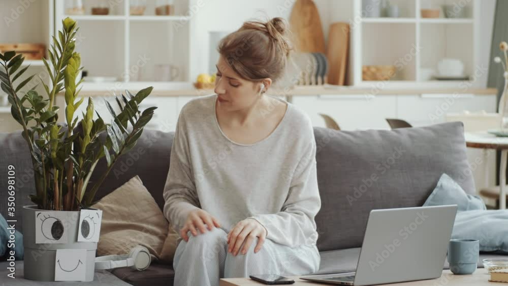 Young bored woman in wireless headphones lying on couch and listening to music with her imaginary friend as houseplant while staying alone at home under lockdown Young bored woman in wireless headphones lying on couch and listening to music with her imaginary friend as houseplant while staying alone at home under lockdown