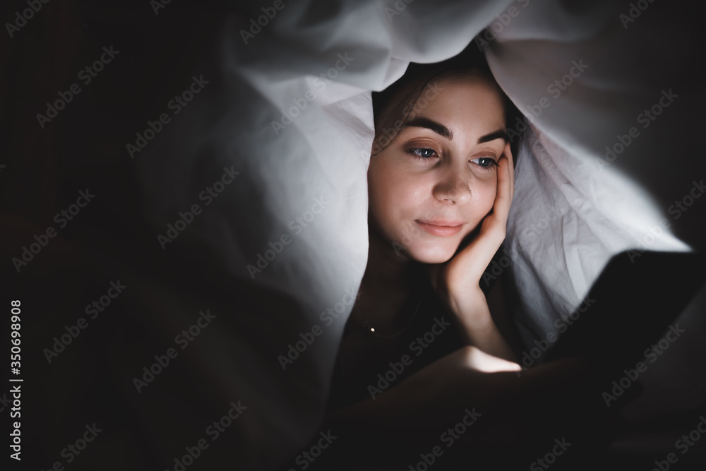 Young smiling woman uses a smartphone, lies under a blanket on a bed at home in the evening. Young smiling woman uses a smartphone, lies under a blanket on a bed at home in the evening.