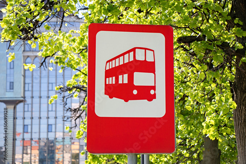 Red bus stop sign for red double decker bus against the background of green foliage in summer on sunny day