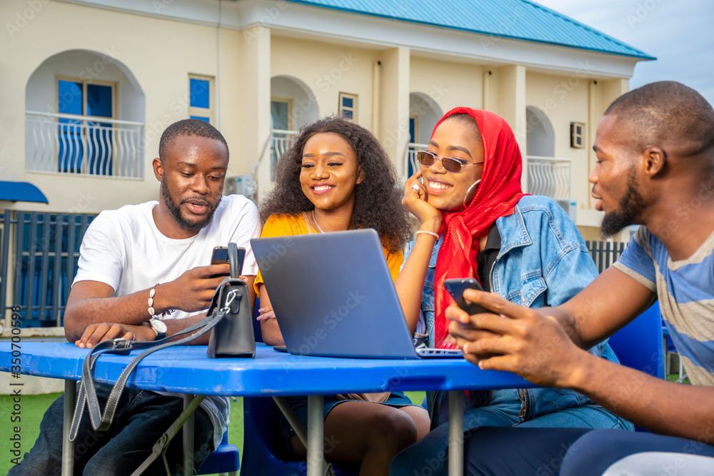 Obraz premium group of young black friends hanging out outdoors, smiling, having a good time, using phones and a laptop computer