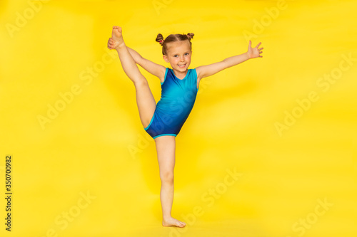 Young cute barefooted gymnast girl with two buns in blue sport suit posing on yellow background with copy space.