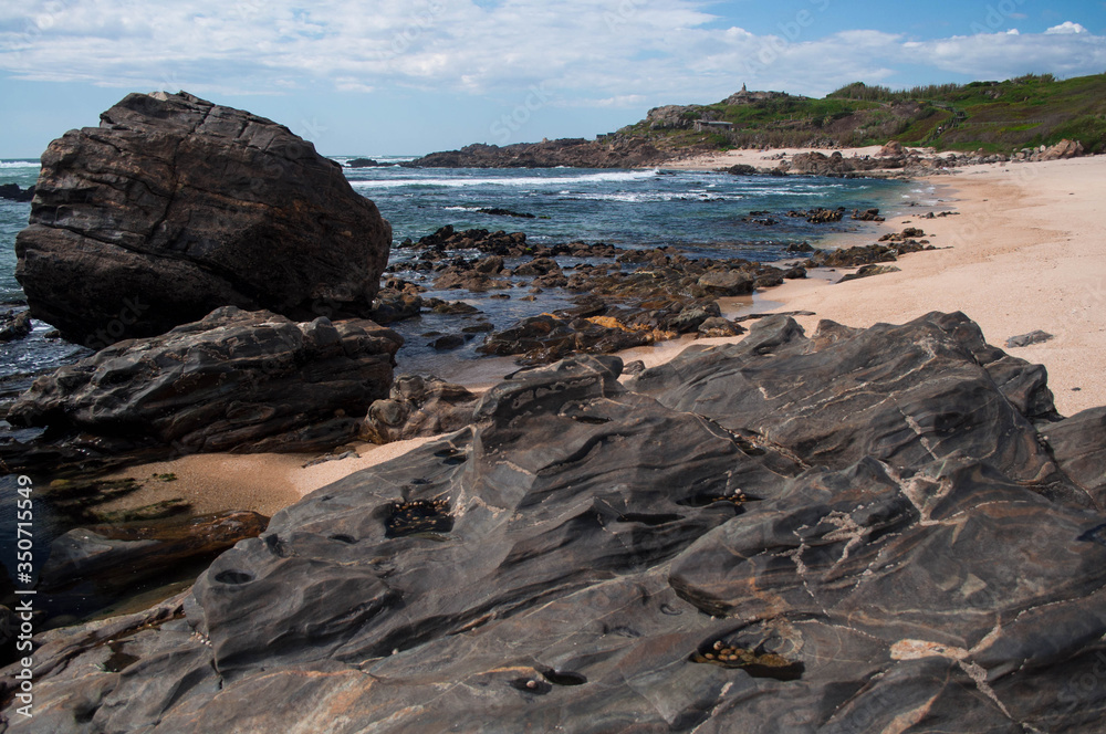 A rocky beach with brown stones, which have been influenced by azul waves over the decades in the foreground. In the background, the hill is covered with green grass