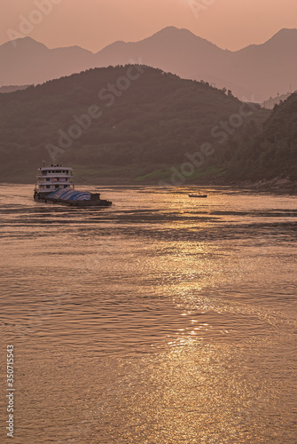Chongqing, China - May 8, 2010: Evening light on Yangtze River. Loaded barge sails on while golden sunset reflected by brown water in front of shoreline with forested hills. Small fishing sloop presen