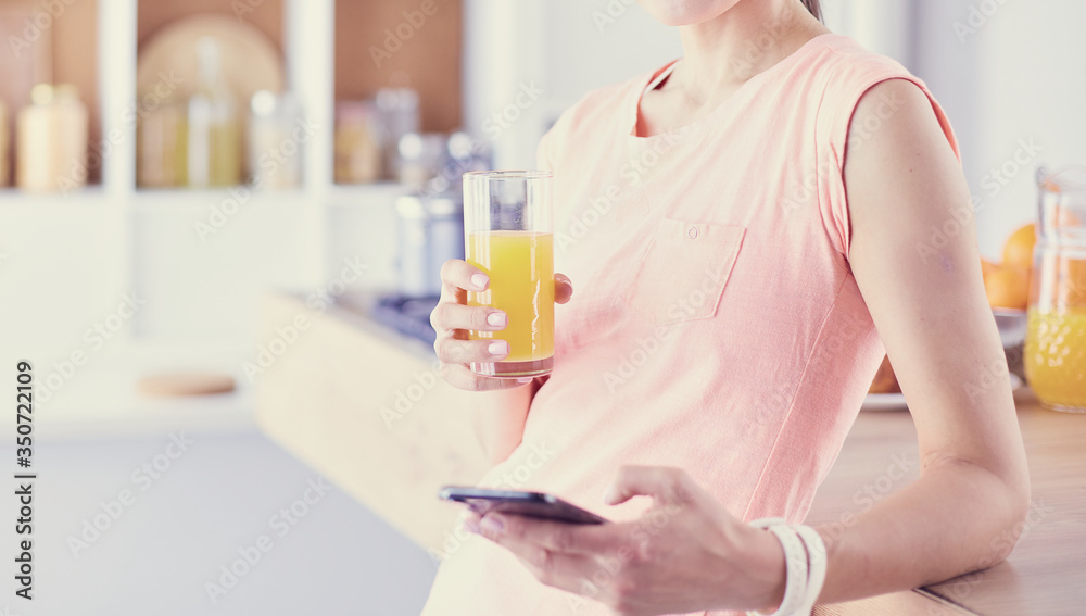 Smiling pretty woman looking at mobile phone and holding glass of orange juice while having breakfast in a kitchen. Smiling pretty woman looking at mobile phone and holding glass of orange juice while having breakfast in a kitchen.