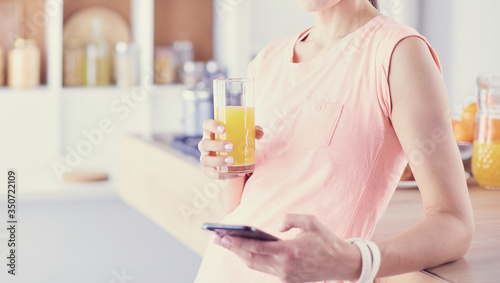 Smiling pretty woman looking at mobile phone and holding glass of orange juice while having breakfast in a kitchen.