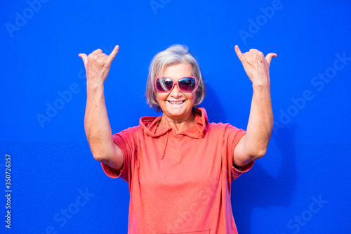 close up and portrait of funny and crazy senior or mature woman doing surfing sign and smililing looking at the camera with blue background - pensioner wearing red clothes and sunglasses happy people